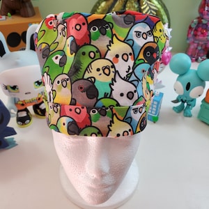 May include: A colorful chef's hat featuring a repeating pattern of various parrots in shades of green, yellow, red, and blue. The hat is displayed on a white mannequin head, showcasing its design and shape. The hat is made of fabric.