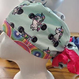 May include: A turquoise blue surgical scrub hat with a rainbow trim and a repeating pattern of unicorns lifting weights. The hat has a pink tie.