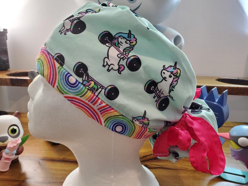 May include: A turquoise blue surgical scrub hat with a rainbow trim and a repeating pattern of unicorns lifting weights. The hat has a pink tie.
