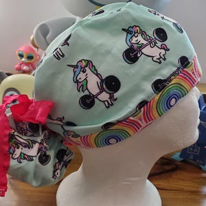 May include: A turquoise blue surgical scrub hat with a rainbow trim. The hat features a repeating pattern of white unicorns lifting pink dumbbells.