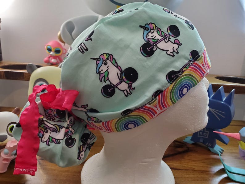 May include: A turquoise blue surgical scrub hat with a rainbow trim. The hat features a repeating pattern of white unicorns lifting pink dumbbells.