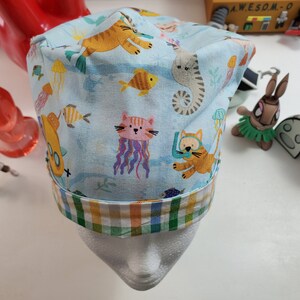 May include: A blue and white patterned scrub hat with a green, yellow, and orange plaid trim. The pattern features cartoon cats, fish, and seahorses.