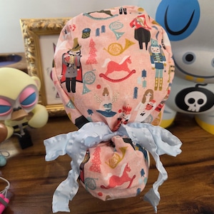 May include: A pink patterned scrub cap featuring a repeating design of toy soldiers, rocking horses, and musical instruments. The cap is tied with a light blue ribbon. The cap is likely for medical professionals.