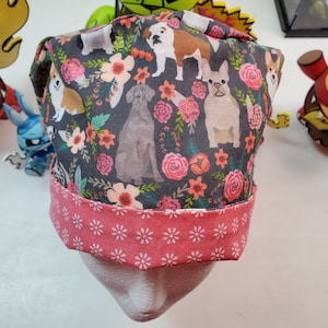 May include: A pink and gray scrub hat with a floral pattern and various dog breeds printed on it. The hat has a pink and white patterned band around the bottom.