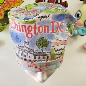 May include: A white fabric headband with a colorful print of Washington D.C. landmarks, including the Capitol Building, the White House, and the National Mall. The headband has a floral print on the bottom edge.