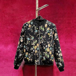 May include: Black bomber jacket with a floral pattern in shades of yellow, blue, and green. The jacket has a black collar, cuffs, and hem, and a silver zipper closure. The jacket is hanging on a black hanger.