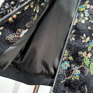 May include: Black jacket with floral embroidery in shades of blue, green, and orange. The jacket features a silver zipper and a black lining. The floral pattern is detailed and covers the jacket's exterior.