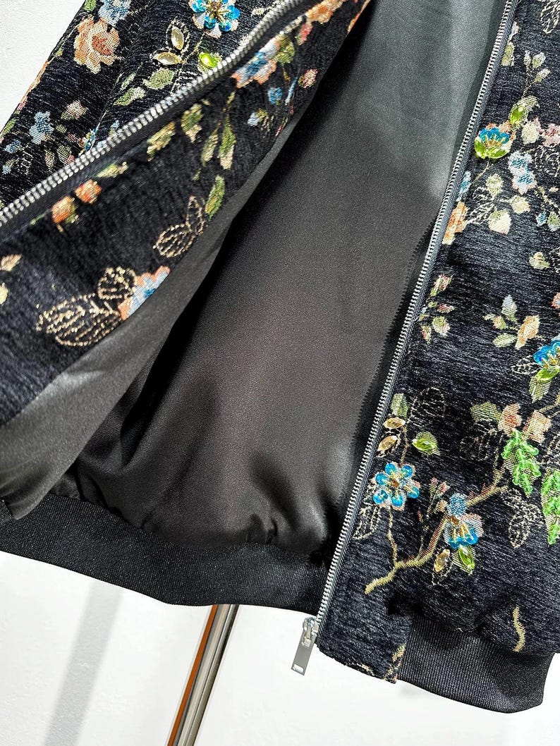 May include: Black jacket with floral embroidery in shades of blue, green, and orange. The jacket features a silver zipper and a black lining. The floral pattern is detailed and covers the jacket's exterior.
