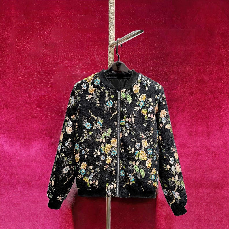 May include: Black bomber jacket with a floral pattern in shades of yellow, blue, and green. The jacket has a black collar, cuffs, and hem, and a silver zipper closure. The jacket is hanging on a black hanger.