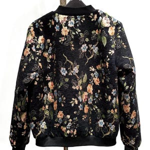 May include: Black bomber jacket with a floral pattern in shades of peach, blue, and cream. The jacket has a black collar, cuffs, and hem. The floral design covers the entire jacket, creating a vintage aesthetic.