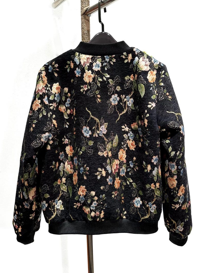 May include: Black bomber jacket with a floral pattern in shades of peach, blue, and cream. The jacket has a black collar, cuffs, and hem. The floral design covers the entire jacket, creating a vintage aesthetic.