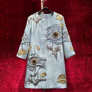 May include: A pale blue coat with floral embroidery. The coat features large, detailed flower designs in shades of grey, yellow, and black. The coat has three-quarter sleeves and is hanging on a black hanger.