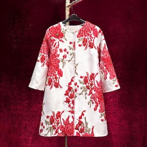 May include: A white coat featuring a vibrant red floral pattern with intricate embroidery and beadwork. The garment has a round neckline, three-quarter length sleeves, and a straight silhouette, hanging on a black hanger against a dark red background.