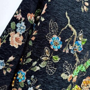 May include: Close-up of a dark fabric with an embroidered floral pattern. The design features colorful flowers in shades of blue, pink, and gold, with green leaves and stems. The fabric has a textured appearance, and the embroidery adds a touch of elegance.
