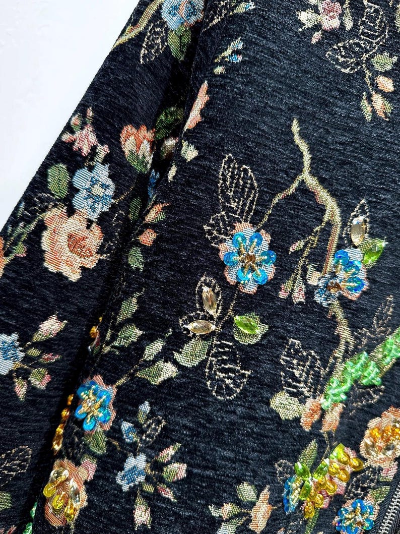May include: Close-up of a dark fabric with an embroidered floral pattern. The design features colorful flowers in shades of blue, pink, and gold, with green leaves and stems. The fabric has a textured appearance, and the embroidery adds a touch of elegance.