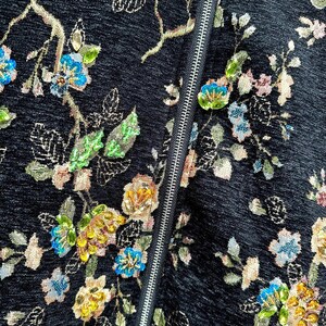 May include: Close-up of a black fabric with an embroidered floral pattern. The design features colorful flowers in shades of blue, yellow, and pink, with green leaves and gold accents. A silver zipper runs vertically down the center.