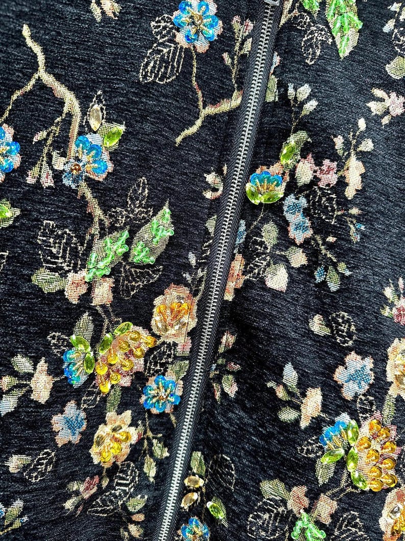 May include: Close-up of a black fabric with an embroidered floral pattern. The design features colorful flowers in shades of blue, yellow, and pink, with green leaves and gold accents. A silver zipper runs vertically down the center.