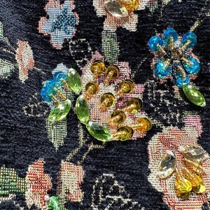 May include: Close-up of a black fabric with embroidered floral patterns. The design features various flowers in shades of pink, blue, and gold, embellished with clear and gold-colored beads. The fabric has a textured appearance.
