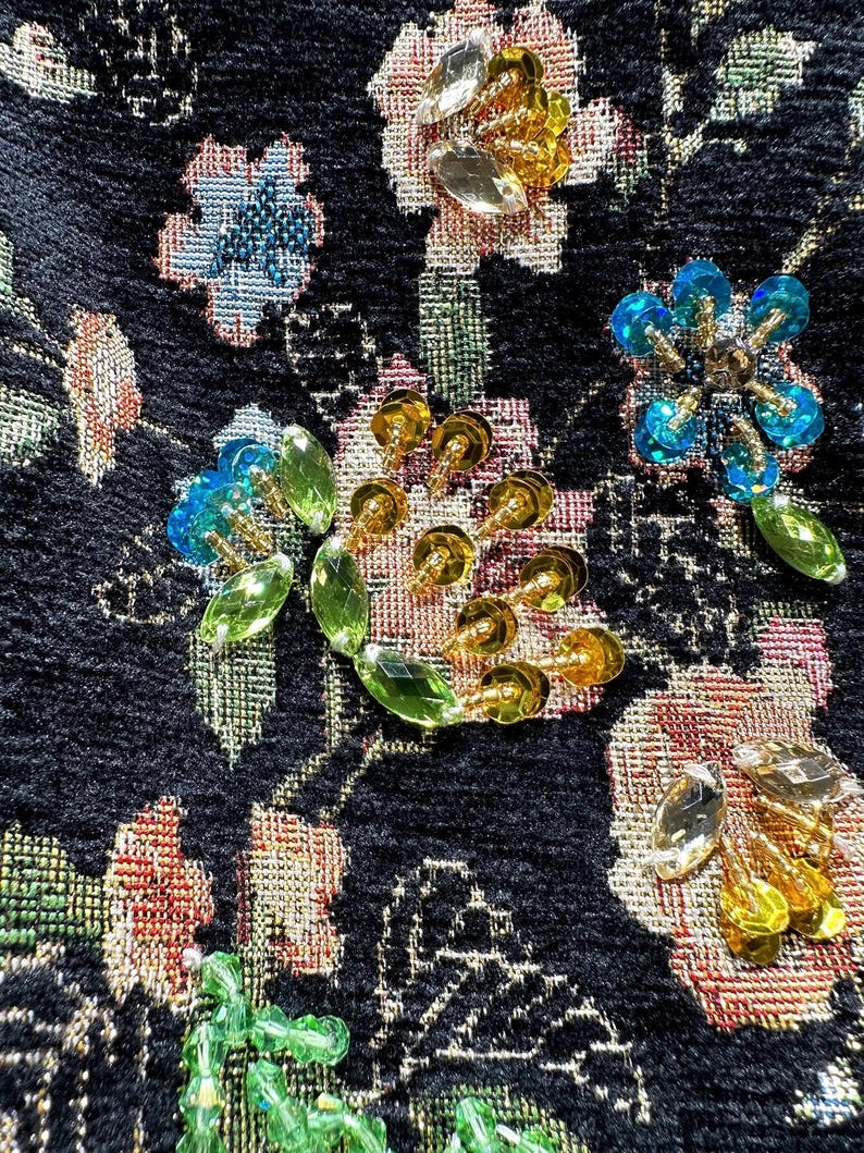 May include: Close-up of a black fabric with embroidered floral patterns. The design features various flowers in shades of pink, blue, and gold, embellished with clear and gold-colored beads. The fabric has a textured appearance.