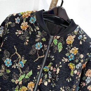 May include: Black bomber jacket with a floral pattern. The jacket features a black ribbed collar and cuffs, a front zipper, and a colorful embroidered floral design in shades of blue, yellow, and green.