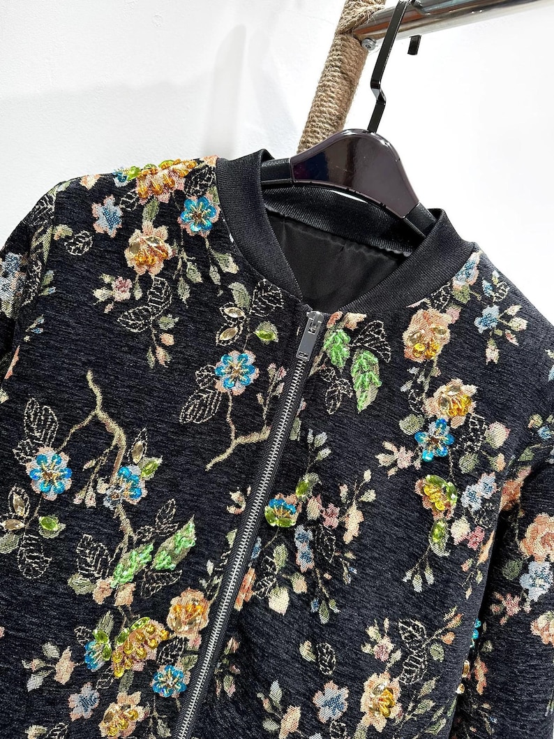 May include: Black bomber jacket with a floral pattern. The jacket features a black ribbed collar and cuffs, a front zipper, and a colorful embroidered floral design in shades of blue, yellow, and green.