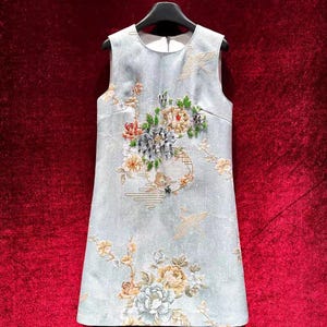 Peut inclure: Robe sans manches bleu clair avec broderie florale. La robe a un col rond et une coupe droite. La broderie comprend des fleurs roses, jaunes et vertes, avec des détails dorés. La robe est suspendue à un cintre noir.