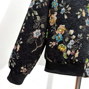 May include: Black jacket with floral embroidery. The jacket features a black ribbed cuff and hem. The floral design includes pink, blue, and yellow flowers with green leaves. The jacket has a textured appearance.
