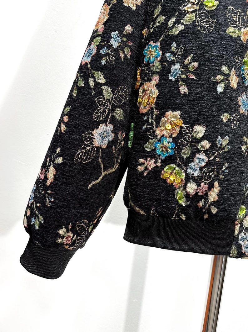 May include: Black jacket with floral embroidery. The jacket features a black ribbed cuff and hem. The floral design includes pink, blue, and yellow flowers with green leaves. The jacket has a textured appearance.