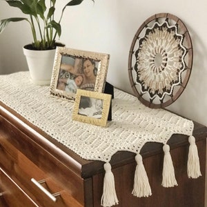May include: A white crocheted table runner with tassels on a wooden dresser. The runner is decorated with two framed photos and a macrame wall hanging.