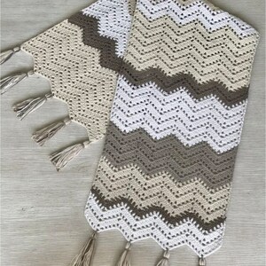 May include: A crocheted table runner with a chevron pattern in beige, brown, and white. The runner has tassels on each end.