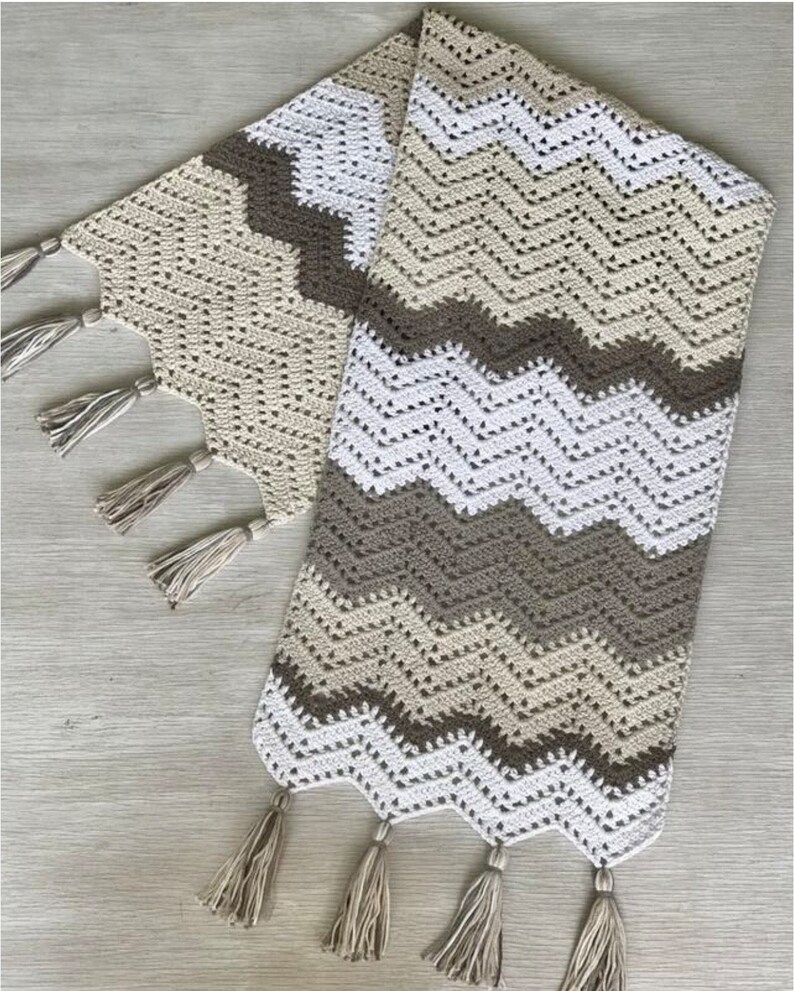 May include: A crocheted table runner with a chevron pattern in beige, brown, and white. The runner has tassels on each end.