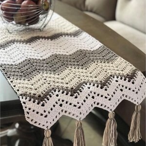 May include: A crocheted table runner with a white, beige, and brown chevron pattern. The runner has a scalloped edge and tassels on the ends.