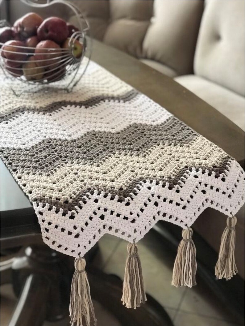 May include: A crocheted table runner with a white, beige, and brown chevron pattern. The runner has a scalloped edge and tassels on the ends.