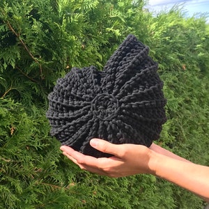 May include: A dark charcoal grey crocheted cushion shaped like a nautilus shell. The item features a dense, textured yarn construction with a spiralling pattern that radiates from a central circular point, creating a distinct, rounded, and ribbed form.