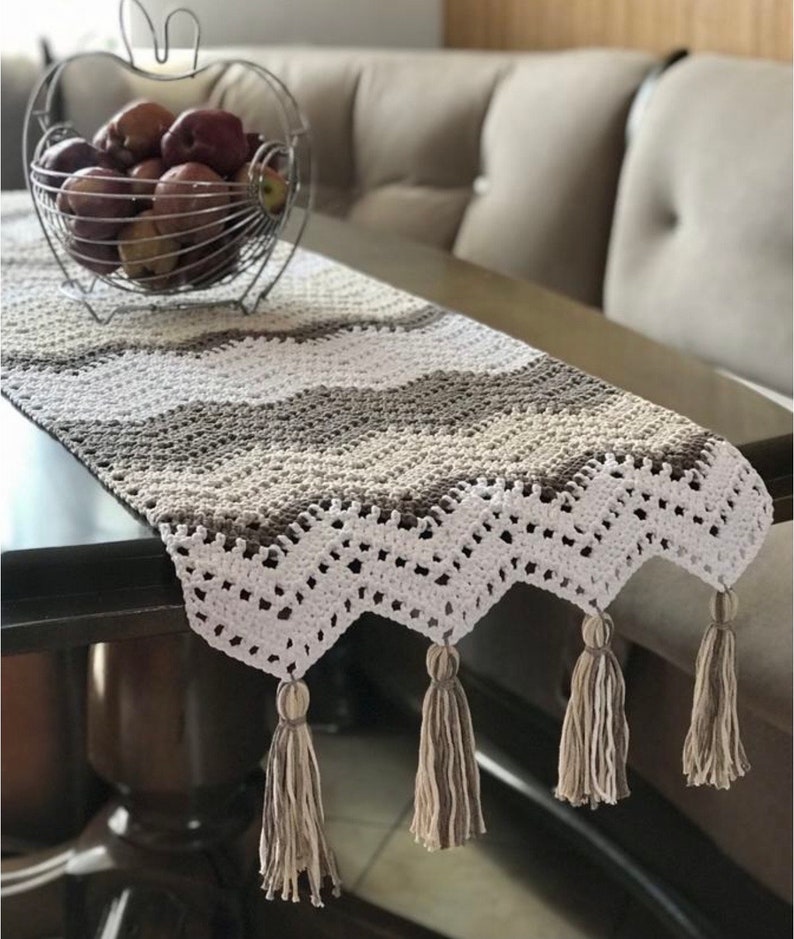 May include: A crocheted table runner with a chevron pattern in white, beige, and brown. The runner has tassels on the ends.