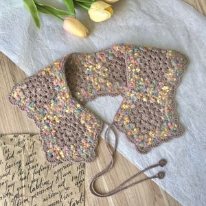 Op de afbeelding: Een bruin gehaakt kraagje met een kleurrijk gespikkeld patroon en een strik sluiting. De kraag ligt op een wit oppervlak met een stuk papier met handgeschreven tekst.