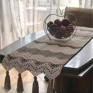 May include: A crocheted table runner with a white and brown chevron pattern and tassels. The runner is on a wooden table with a metal apple-shaped fruit bowl filled with apples.