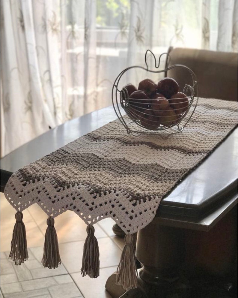May include: A crocheted table runner with a white and brown chevron pattern and tassels. The runner is on a wooden table with a metal apple-shaped fruit bowl filled with apples.