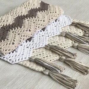 May include: A crocheted throw blanket with a chevron pattern in beige, brown, and white. The blanket has fringe tassels in beige and white.