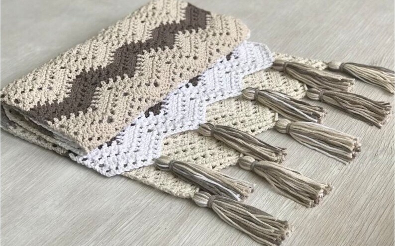 May include: A crocheted throw blanket with a chevron pattern in beige, brown, and white. The blanket has fringe tassels in beige and white.