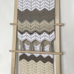 May include: A crocheted throw blanket with a chevron pattern in white, beige, and brown. The blanket has fringe on the bottom and is hanging on a wooden ladder.
