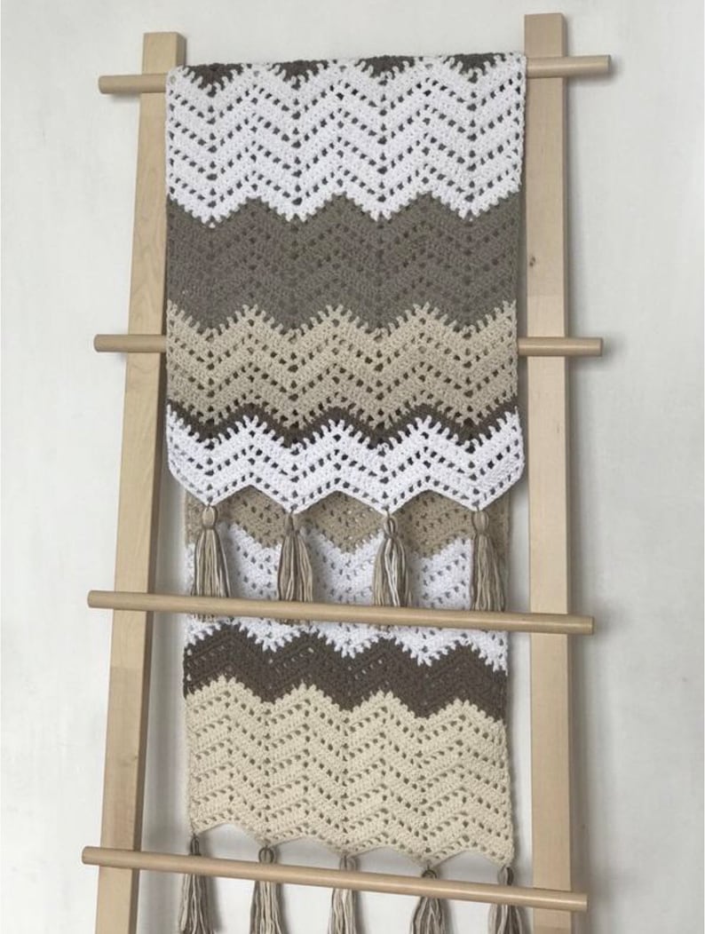 May include: A crocheted throw blanket with a chevron pattern in white, beige, and brown. The blanket has fringe on the bottom and is hanging on a wooden ladder.