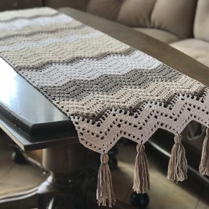 May include: A crocheted table runner with a chevron pattern in shades of brown, beige, and white. The runner has a delicate lace trim and tassels on the ends.