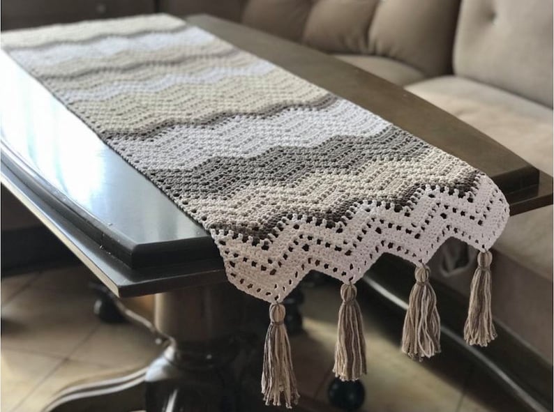 May include: A crocheted table runner with a chevron pattern in shades of brown, beige, and white. The runner has a delicate lace trim and tassels on the ends.