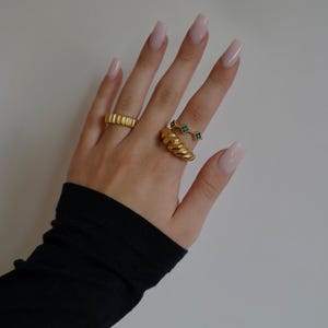 Könnte beinhalten: Eine Hand trägt drei goldene Ringe. Ein Ring ist ein dickes, strukturiertes Band. Ein weiterer Ring ist ein dünnes Band mit einer Reihe kleiner grüner Steine. Der dritte Ring ist ein dünnes Band mit einem einzigen grünen Stein.