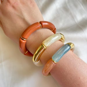 Può includere: Tre braccialetti elastici con perline cilindriche in arancione bruciato tinta unita, crema marmorizzato e azzurro traslucido, intervallate da distanziatori in metallo color oro lucido. I braccialetti sono indossati insieme, mostrando un mix di texture lisce, lucide e metalliche.