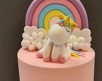 May include: A pink cake decorated with a white unicorn figurine, a rainbow, and white clouds. The unicorn has a gold horn and a yellow mane. The cake is decorated with gold glitter.