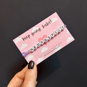 May include: A pink card with white clouds and a pink heart with the text "keep going, babe!" and a white beaded bracelet with the text "NEUROSPICY" in black letters. The bracelet is on a pink background with white clouds and a pink heart. The text "THE URBAN PONYTAIL" and the website "theurbanponytail.etsy.com" are printed on the card.