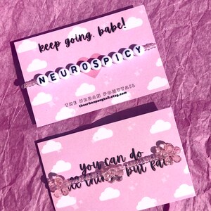 May include: Two cards with pink and white cloud designs. The top card has the text "keep going, babe!" and a beaded bracelet with the text "NEUROSPICY". The bottom card has the text "you can do ne thin but fail" and a beaded bracelet with pink flower-shaped beads.