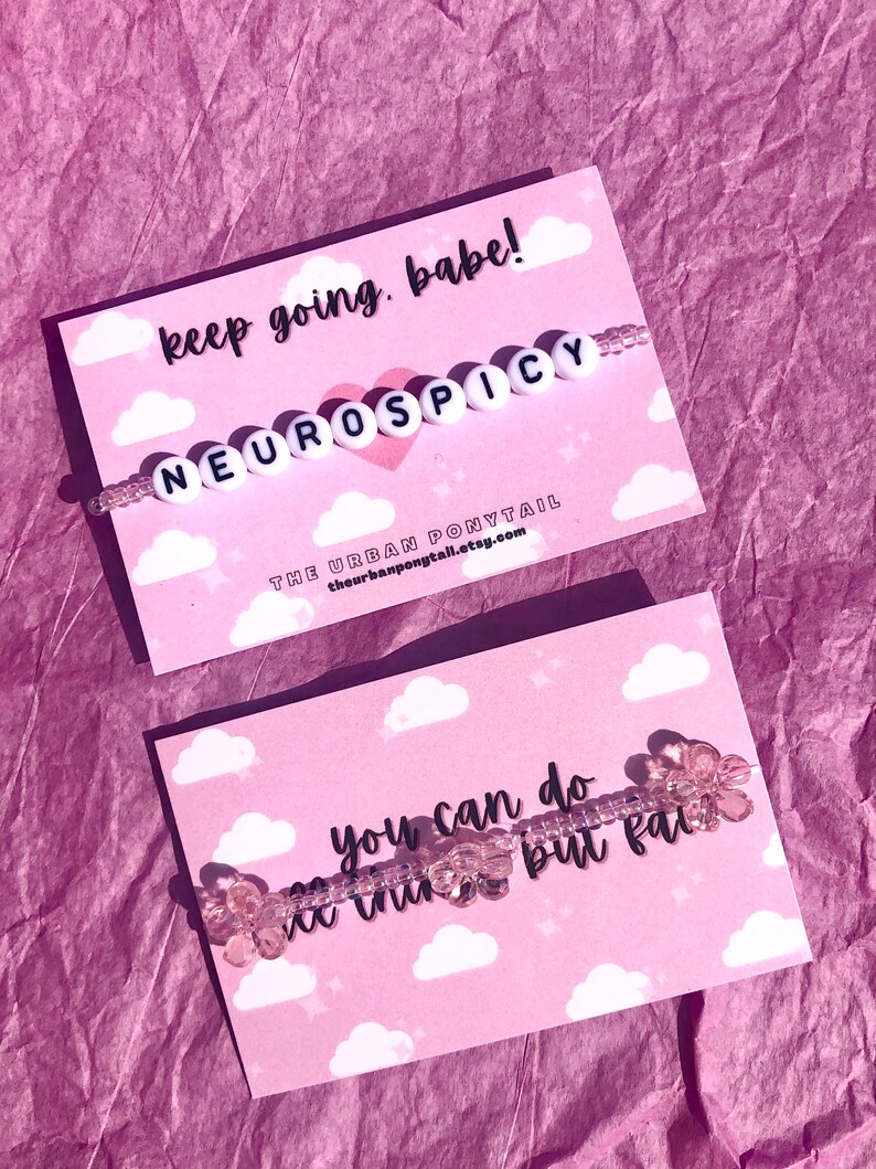 May include: Two cards with pink and white cloud designs. The top card has the text "keep going, babe!" and a beaded bracelet with the text "NEUROSPICY". The bottom card has the text "you can do ne thin but fail" and a beaded bracelet with pink flower-shaped beads.